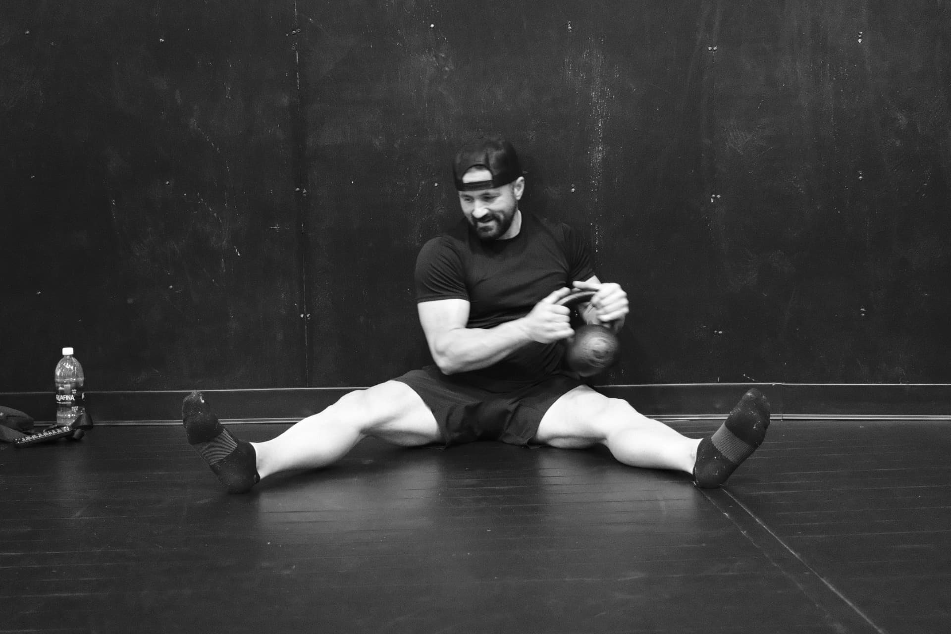 Black and white photo of Shawn Taylor seated in a straddle on the studio floor, smiling and looking down at a kettlebell